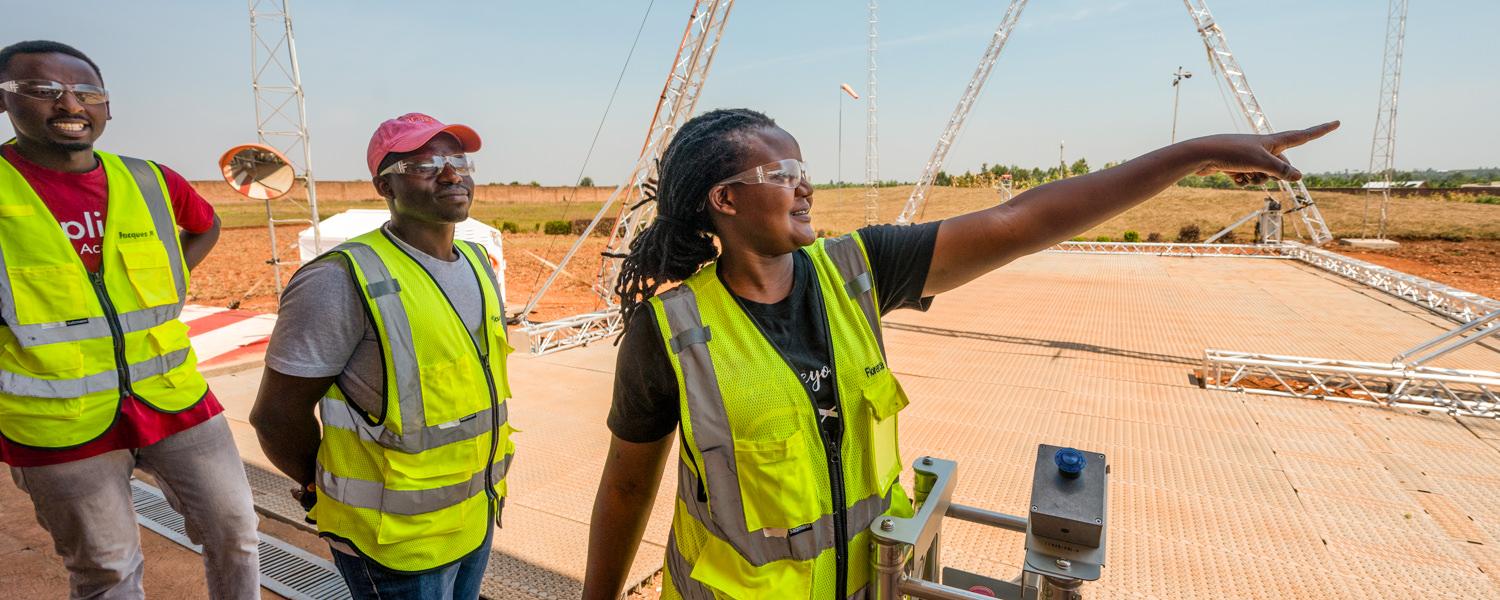 A medium shot of three individuals standing on an outdoor launch pad under a clear sky, surrounded by metal lattice towers. A Rwandan woman in the center, wearing safety glasses and a neon yellow vest, smiles as she points into the distance. Two men in safety vests stand behind her, tracking where she is pointing.