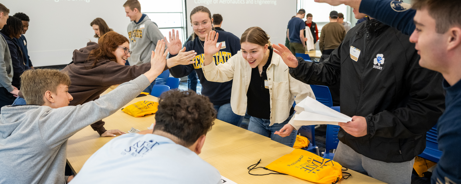 A group of students work around a table in a classroom. Two of the students high five each other.