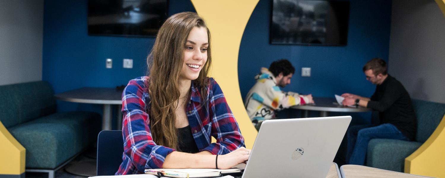 A female student smiling at her laptop with notebooks open beside her