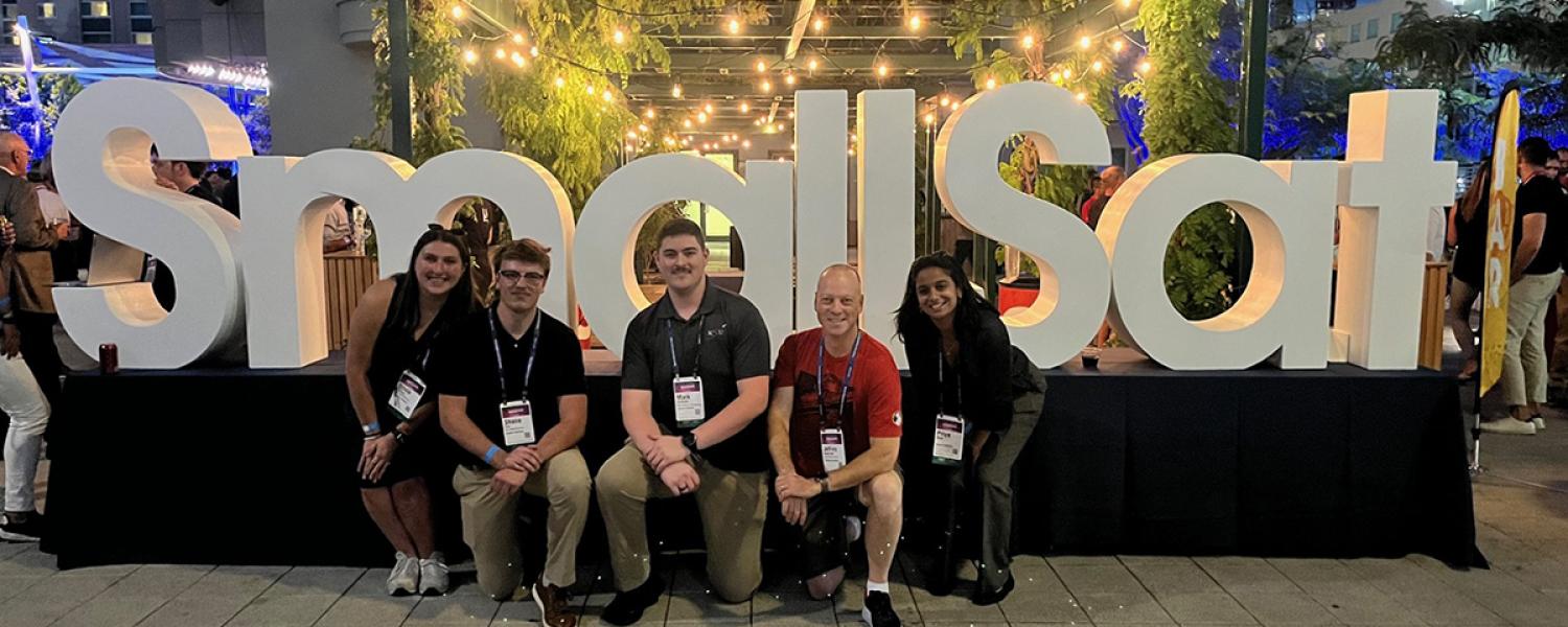 a small group of students standing in front of a large sign that reads "SmallSat"