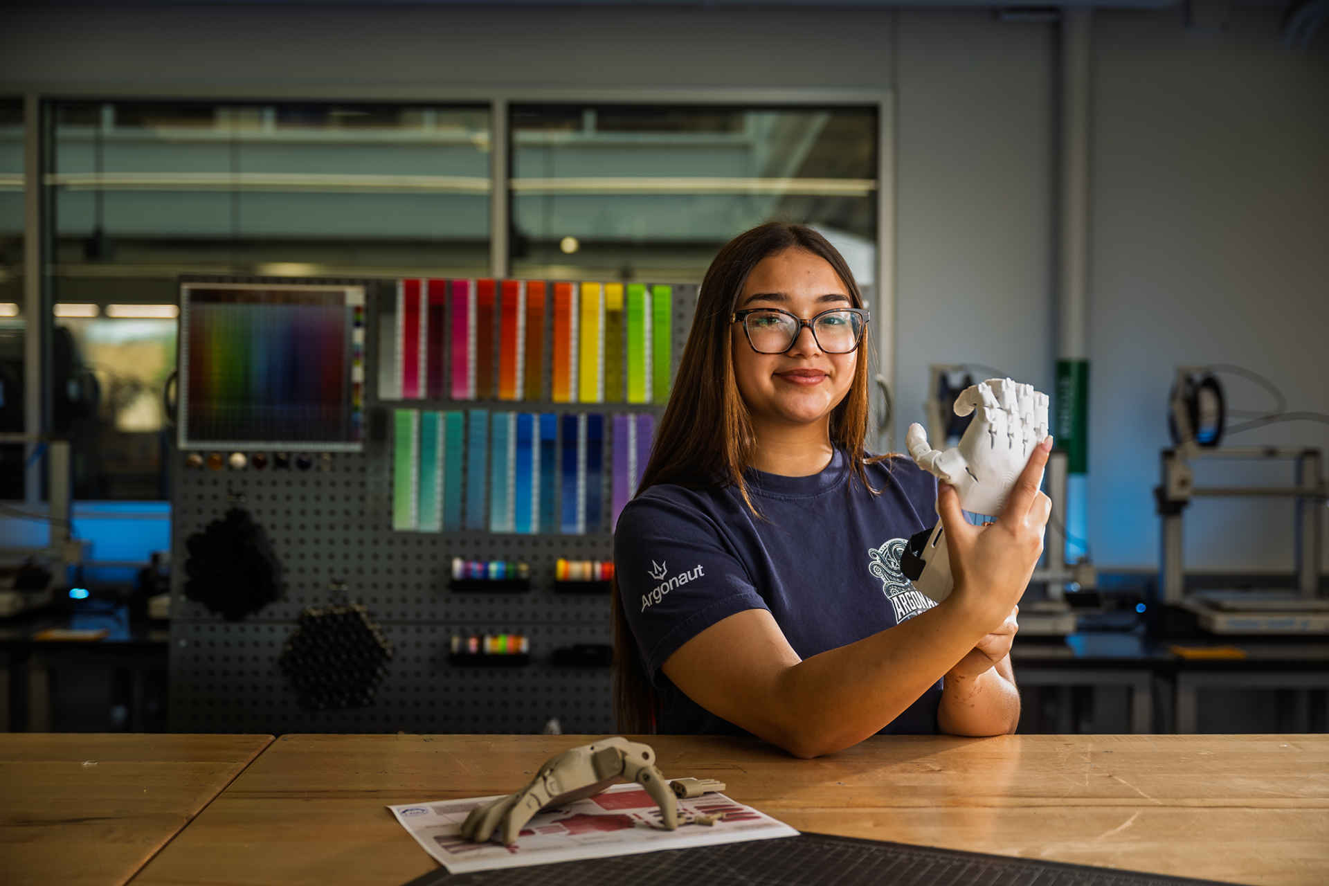 Yariselle Andujar, a ����ɫ��ҹ first-year student wearing an Argonauts t-shirt, holds a white 3D-printed prosthetic hand while seated at a workbench in an engineering lab, with a second prosthetic hand resting on the table in front of her.