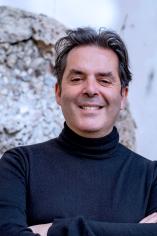 Guido smiles and crosses his arms in a chest-up shot. He wears a black, long-sleeve turtle neck, a similar color to his close-cropped black hair