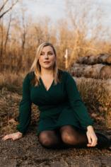 Hannah poses on the forest floor outside, with yellow Fall trees in background. She wears a dark green long-sleeve dress, and has blonde hair
