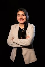 A woman with dark long hair smiles as she crosses her arms and faces the camera, wearing a tan blazer and black top