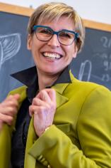 Sabine in a medium shot, clutching her green jacket on either side as she smiles at the camera. Her hair is short and blonde, and she wears gray glasses. 