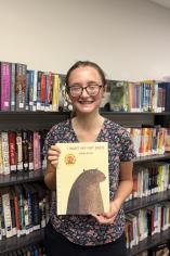 Half-body photograph of Abbigail Thomas holding a book titled "I want my hat back", background consists of a library with books on the shelves