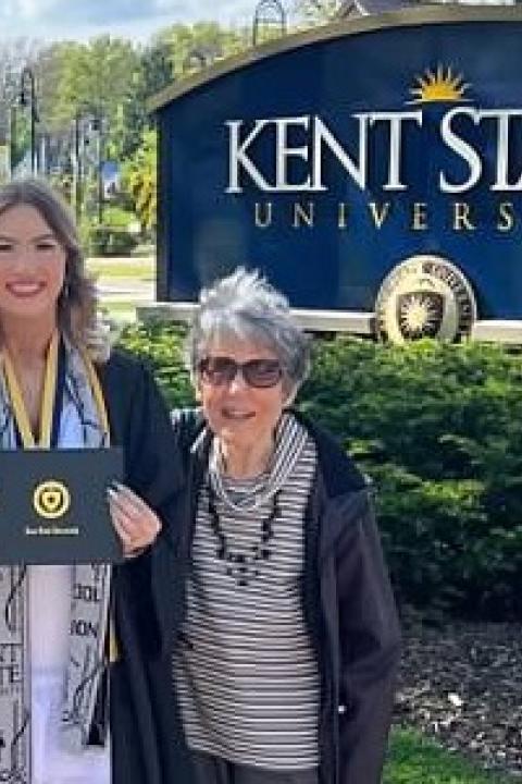 A graduate in cap and gown holding a diploma cover stands with two family members in front of the �Ը������� University sign on a sunny spring day.
