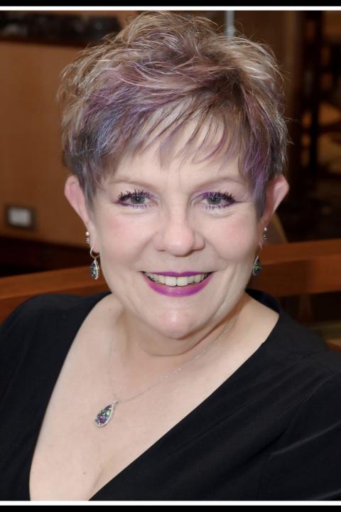Norma West smiling in a professional portrait, wearing a black v-neck top with a teardrop pendant necklace, her short silver hair styled with purple highlights.