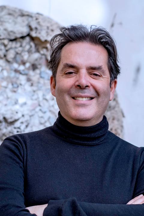 Guido smiles and crosses his arms in a chest-up shot. He wears a black, long-sleeve turtle neck, a similar color to his close-cropped black hair