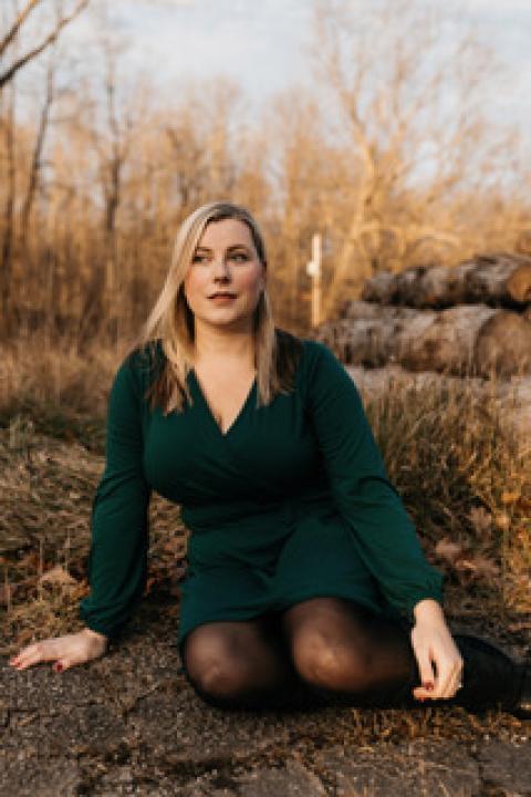 Hannah poses on the forest floor outside, with yellow Fall trees in background. She wears a dark green long-sleeve dress, and has blonde hair