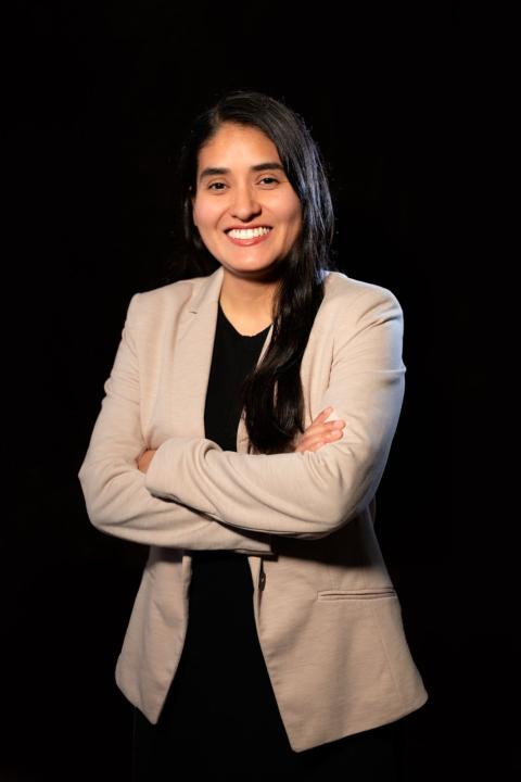 A woman with dark long hair smiles as she crosses her arms and faces the camera, wearing a tan blazer and black top