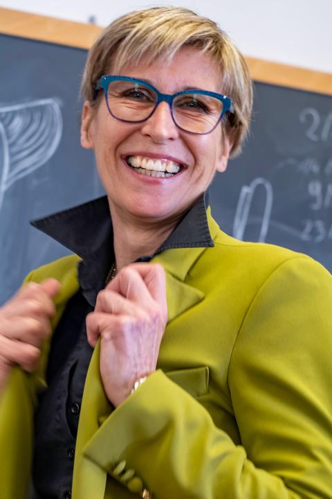 Sabine in a medium shot, clutching her green jacket on either side as she smiles at the camera. Her hair is short and blonde, and she wears gray glasses. 