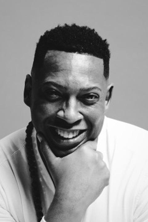 Terance rests his chin on his hand as he smiles at the camera. His hair is dark and close cropped, and he wears a long sleeve white shirt