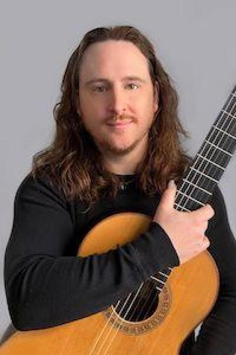 A man with long brown hair holds an acoustic guitar across his chest