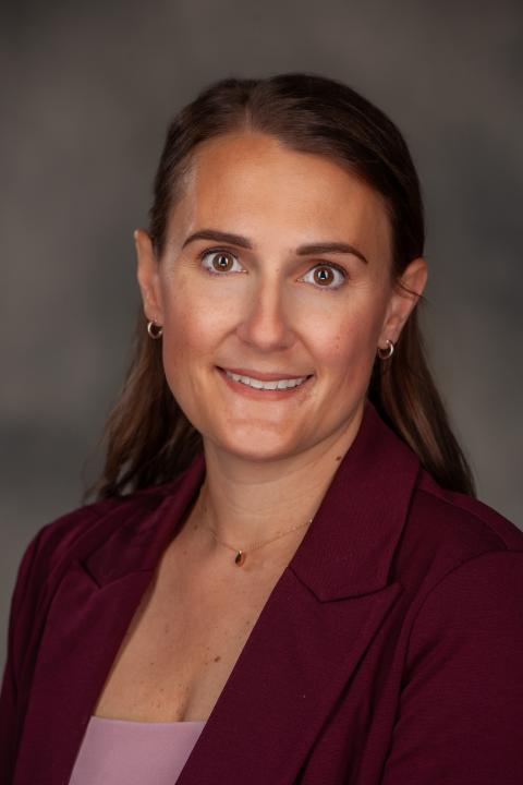 professional headshot of a woman with dark hair half pulled from her face dressed in a dark blazer and pink shirt.