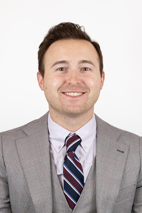 Will Valentini smiling in front of white background. He is wearing a gray suit with a striped tie. 