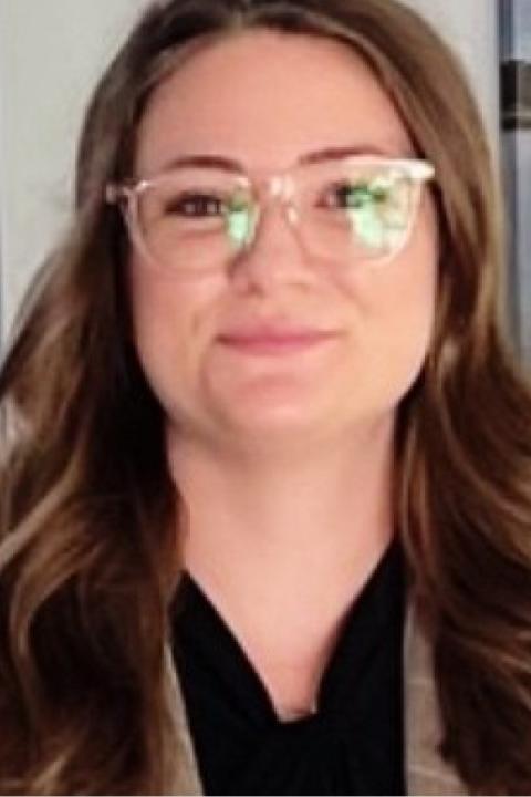 headshot of a woman with long brown hair wearing glasses and a black shirt with a brown blazer on top