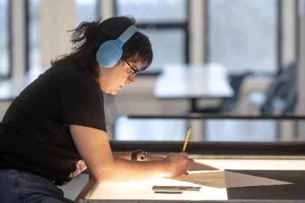 A student wearing headphones sitting at a desk and writing on a piece of paper