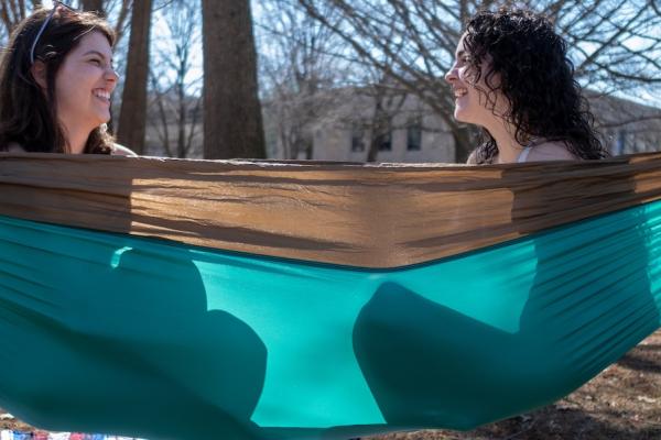 Two students sit in a hammock outside on a sunny day