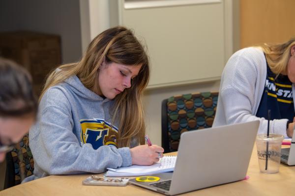 Girl writing on a paper with her computer opened in front of her.