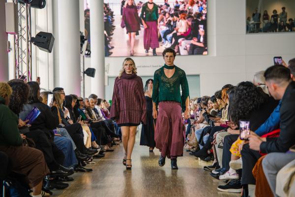 Two models walking down a crowd lined runway inside a building with tall columns and large windows. 