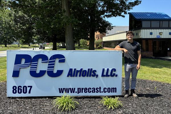 A young man in a t-shirt stands next to a low sign displaying the name of "PCC Airfoils"