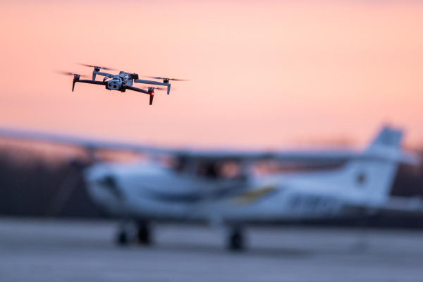 A quadcopter drone flying in the forground with a slightly out of focus small airplane in the background.