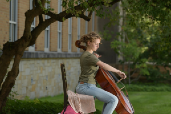 A Kent State cellist playing her cello outside under the shade of a tree