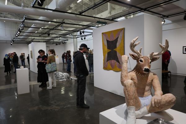Gallery with people looking at art featuring sculpture of a deer head on human body in foreground