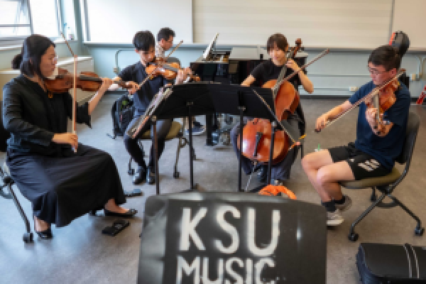 A group of string instrumentalists playing. In the foreground, a music stand that says "KSU Music."