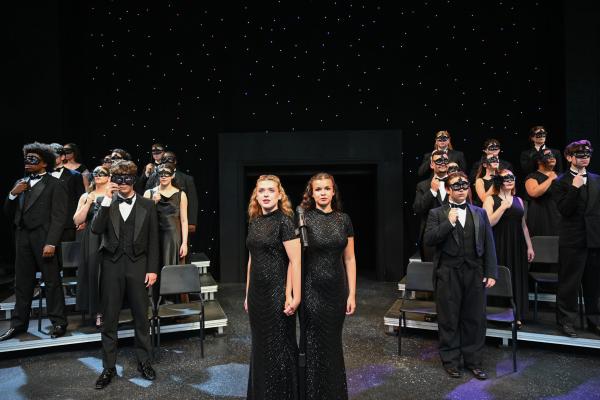 A cast and crew occupy a stage all dressed in black against a starry background. Taking center stage are characters Daisy and Violet, who wear long black dresses and stand conjoined at the hip