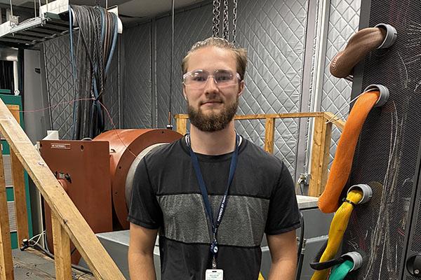A young man wearing a dark t-shirt and jeans in a workroom